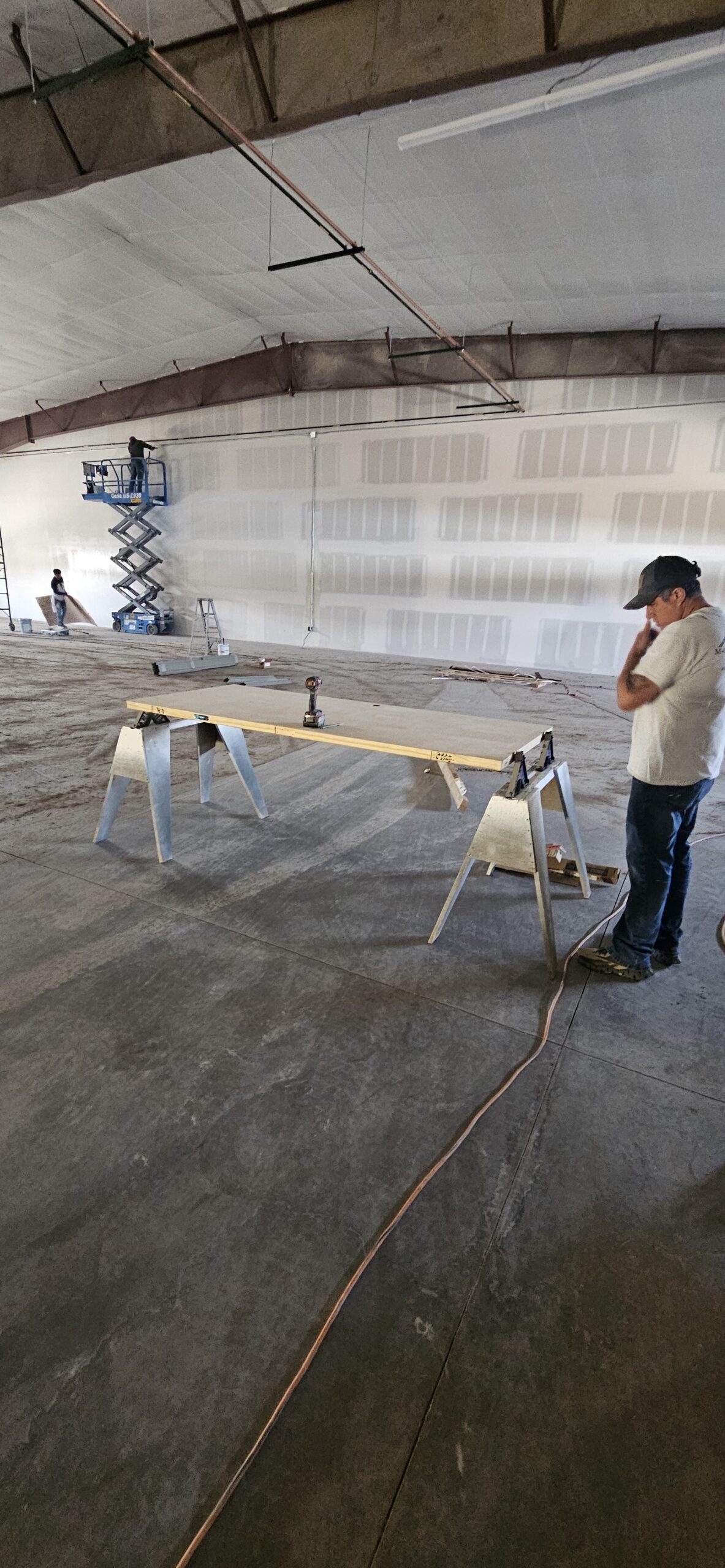 A painting contractor stands by a workbench in a large unfinished warehouse, with construction tools and equipment visible in the background.