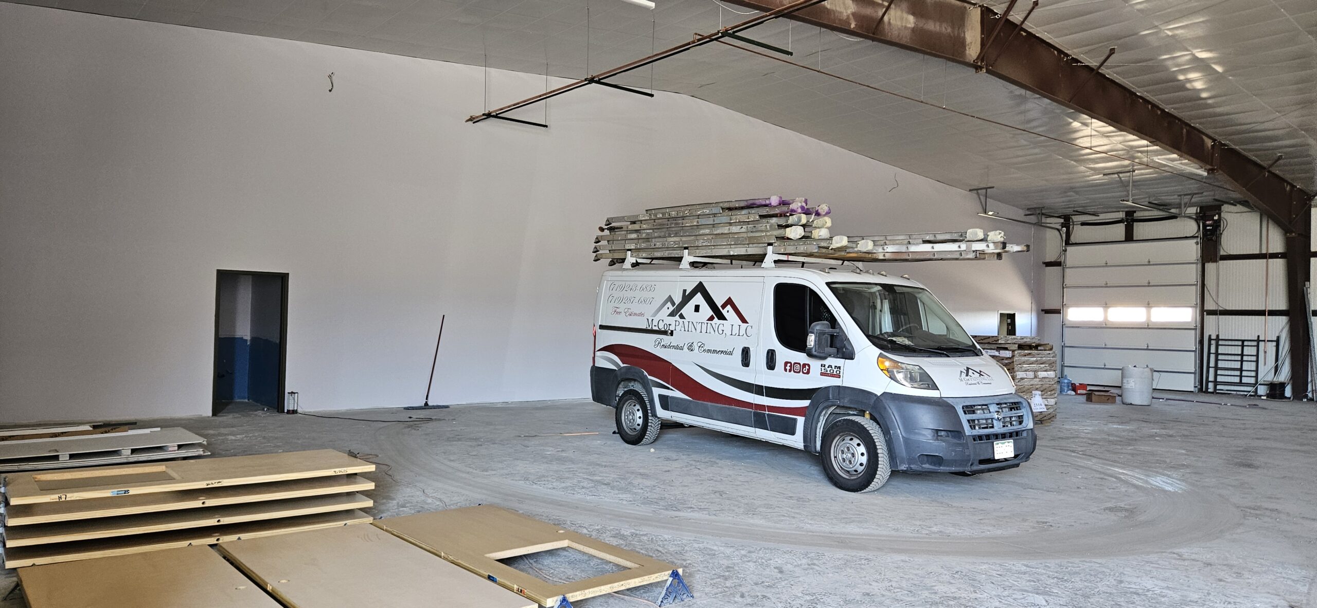 20260319_163514 A white work van with ladders on top, belonging to a painting contractor, is parked inside a large, unfinished industrial building with white walls and scattered construction materials.