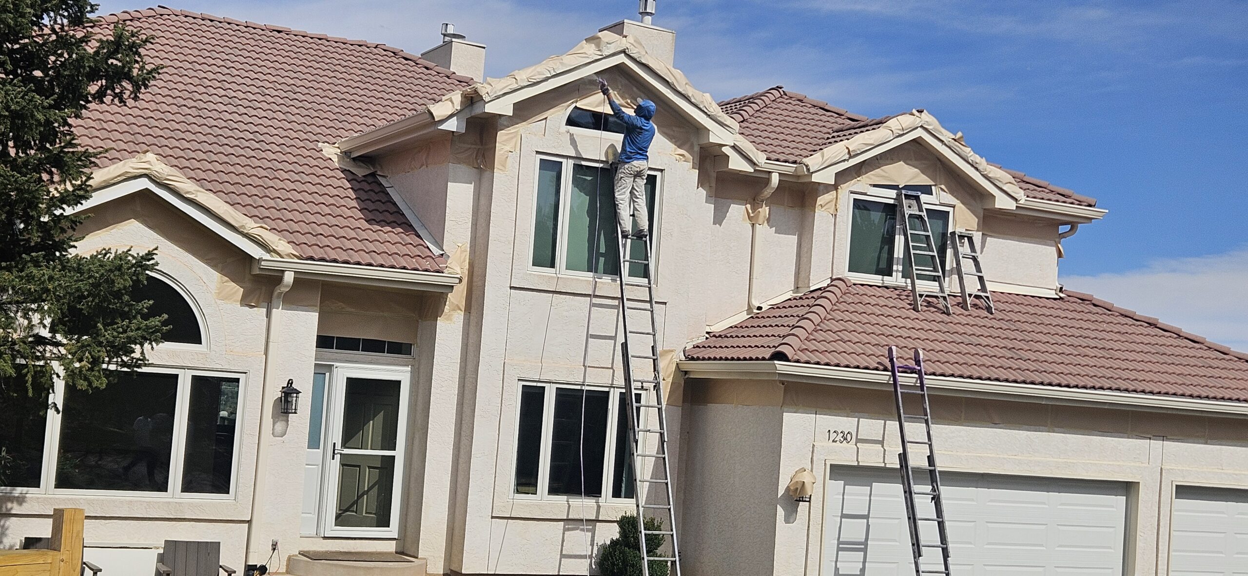 Trim job(3) A painting contractor stands on a tall ladder, carefully painting the exterior trim of a large two-story house with a tile roof. Two other ladders are positioned nearby against the house, ready for additional painters to assist with the work.