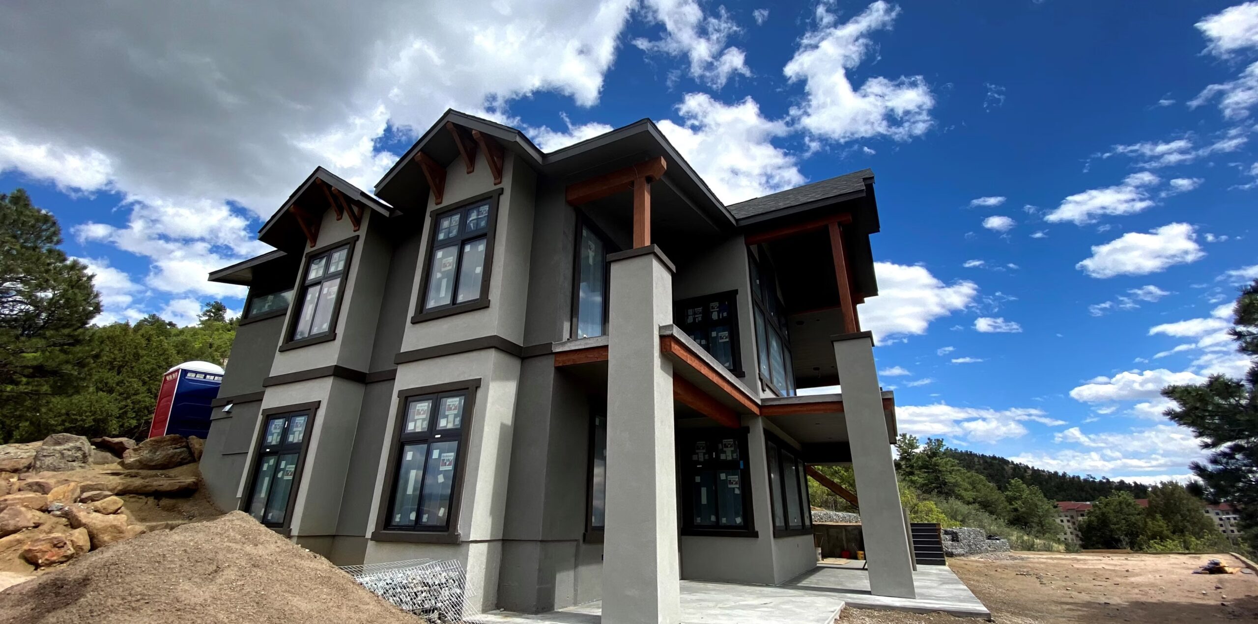 Two-story modern house with large windows and gray stucco exterior, set against a backdrop of trees, rocks, and a blue sky with scattered clouds—an ideal project for a skilled residential painter in Colorado Springs.