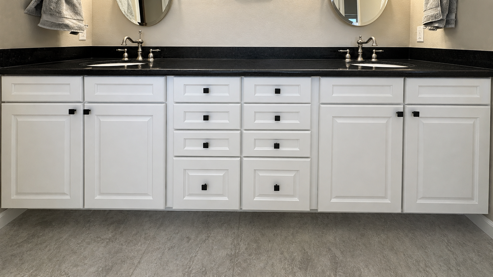 Bathroom Cabinets – Clean White double-sink bathroom vanity with black countertop, chrome faucets, and six drawers between two sets of cabinet doors. Perfect for homeowners seeking a fresh look from a residential painter, this setup features towels hanging on both sides above the sinks—a stylish choice inspired by top painting contractor trends in Colorado Springs.