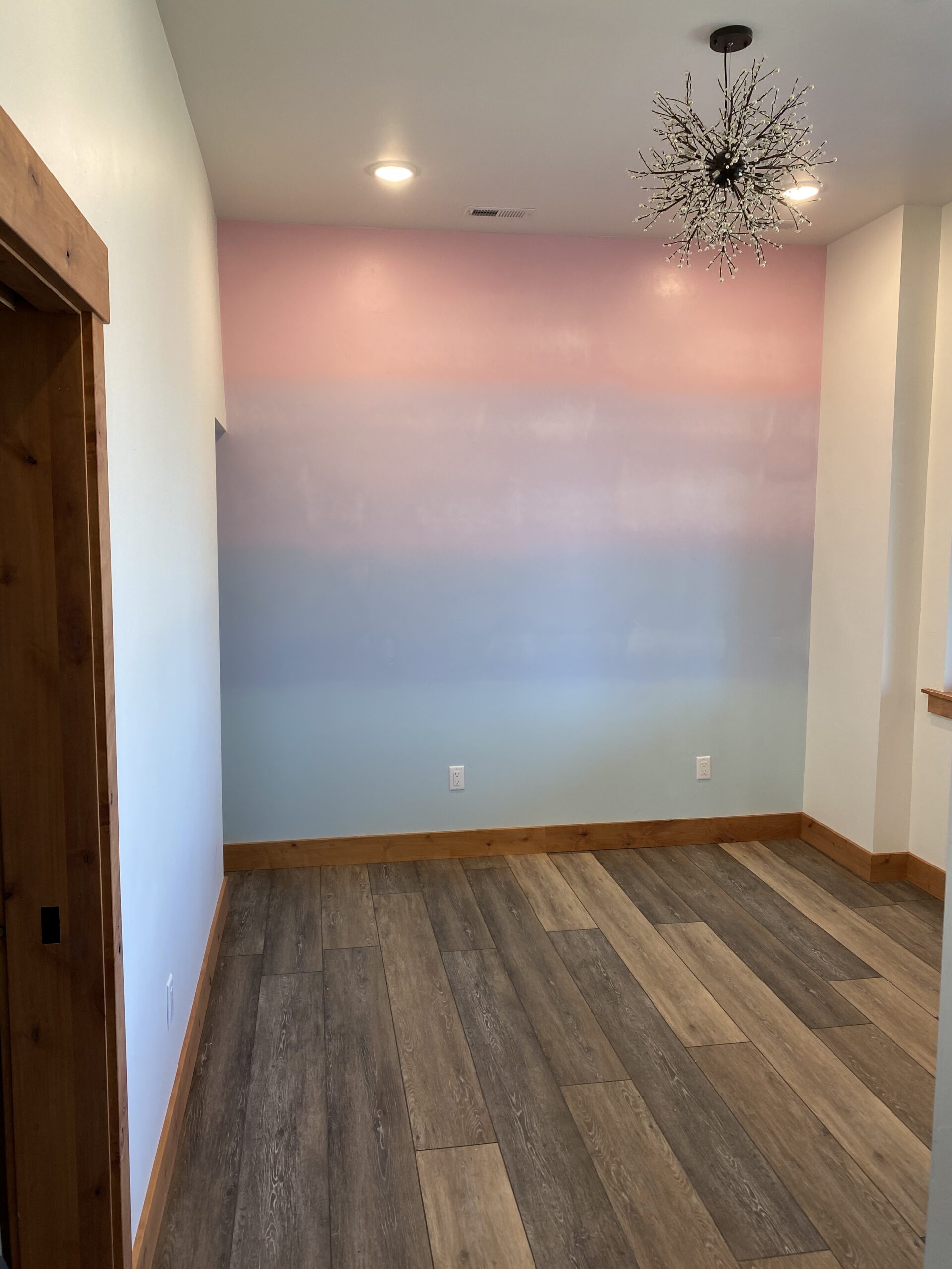 Empty room with wood flooring, a pastel ombre accent wall in pink, purple, and blue, white walls, wood trim, and a modern starburst chandelier on the ceiling—expertly finished by a skilled painting contractor in Colorado Springs.